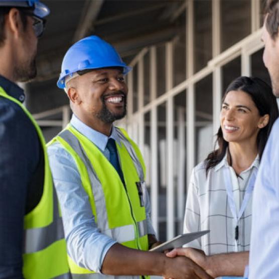 Group of professionals at a construction site, including a smiling man in a blue hard hat and yellow safety vest shaking hands with another person, with other colleagues engaged and appearing happy in the background.