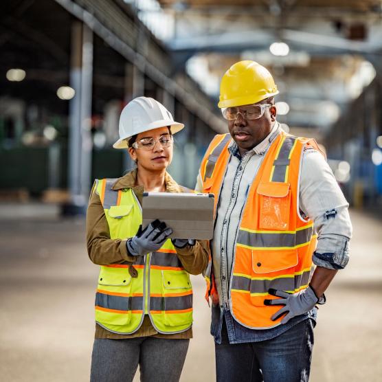 Two construction workers wearing hard hats and safety vests, standing in an industrial setting. One is holding a tablet, and they are both looking at the screen, appearing to discuss or plan something important.